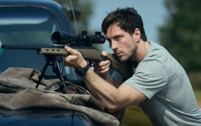 Scenă cu lunetist din thrillerul Fuze (2025) Actor cu pușcă cu lunetă poziționat în spatele unui autoturism, scenă de suspans din filmul Fuze (2025) cu Aaron Taylor-Johnson