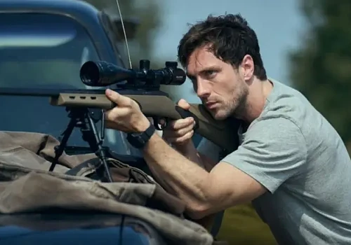 Actor cu pușcă cu lunetă poziționat &icirc;n spatele unui autoturism, scenă de suspans din filmul Fuze (2025) cu Aaron Taylor-Johnson